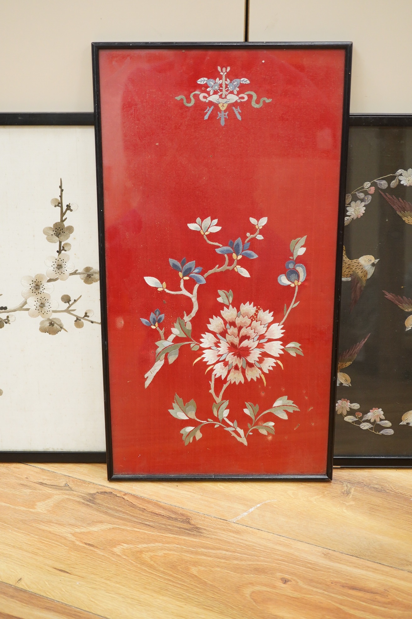 Two Chinese polychromed silk embroidered panels, early 20th century, one on a black silk ground of birds and flowers, the other on a red background of flowers and a monochrome silk embroidered panel of birds and flowers,